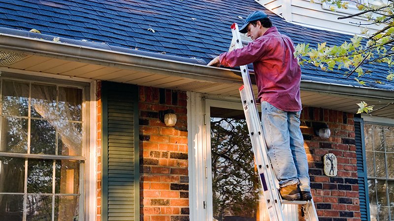 HOME MAINT WEB_001 A person wearing a red shirt and blue cap standing on a ladder, clearing leaves from the gutter of a brick house with white trim and a dark roof.