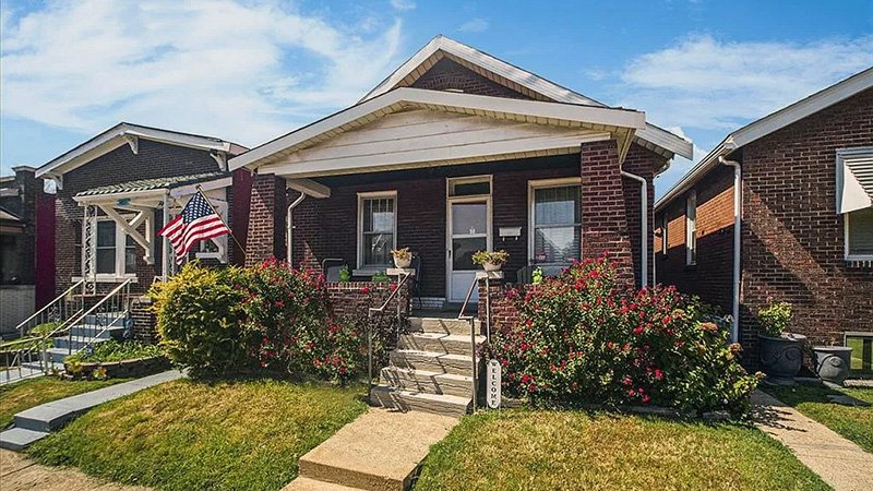 HOMES WEB_001 A single-story brick house with a gabled roof and small front porch, surrounded by a well-kept yard with flowering plants and an American flag, under a sunny sky