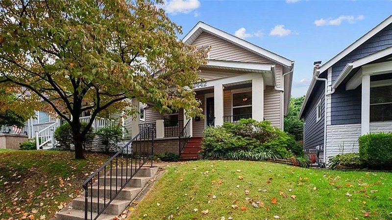 HOMES WEB_002 A suburban house with a gable roof and front porch, set on a grassy lawn with concrete steps and a black railing, surrounded by autumn-colored trees and neighboring homes under a clear sky.
